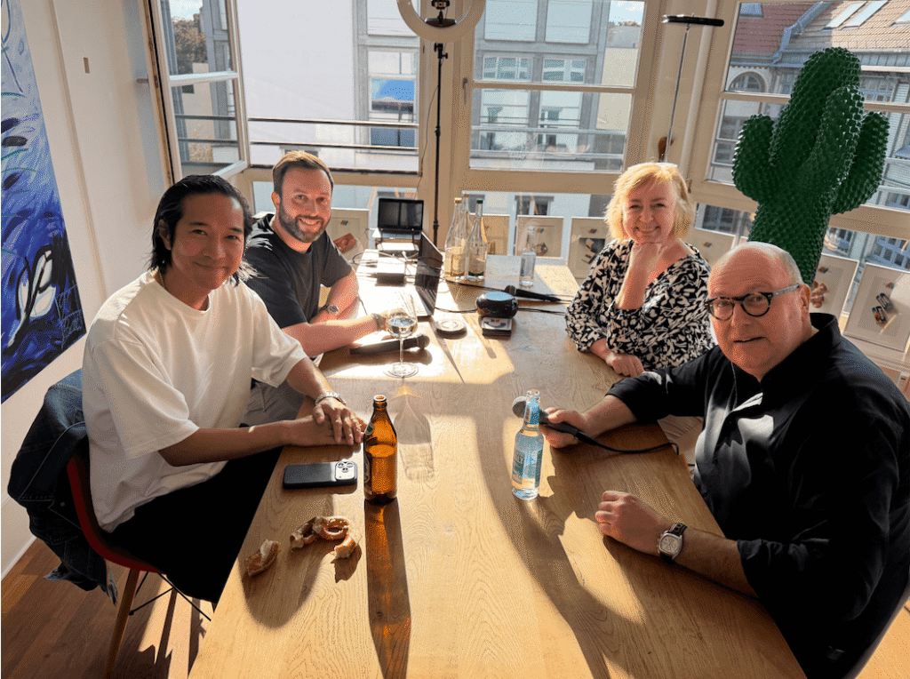 Vier Personen sitzen am Tisch bevor der Podcast losgeht und lächeln in die Kamera.